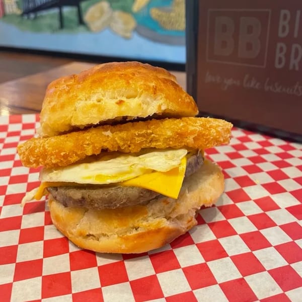 Biscuit Bros' breakfast sandwich with sausage, egg, cheese, and hash brown on red checkered paper.