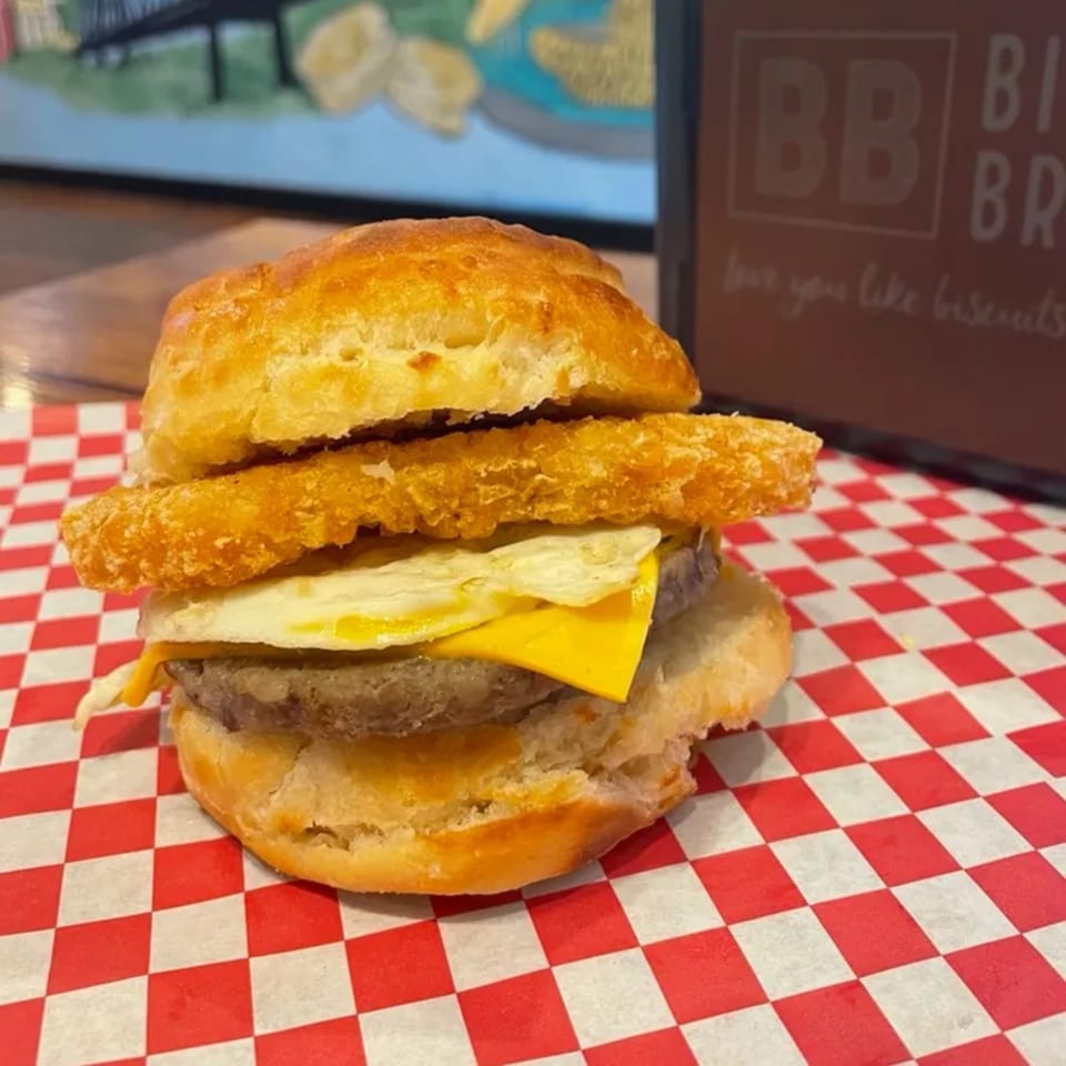 Biscuit Bros' breakfast sandwich with sausage, egg, cheese, and hash brown on red checkered paper.