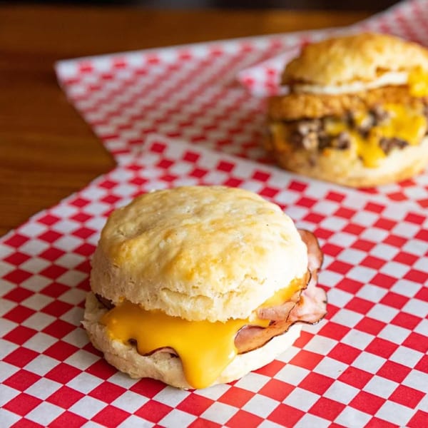 Two Biscuit Bros breakfast sandwiches with melted cheese and meat on red and white checkered paper.