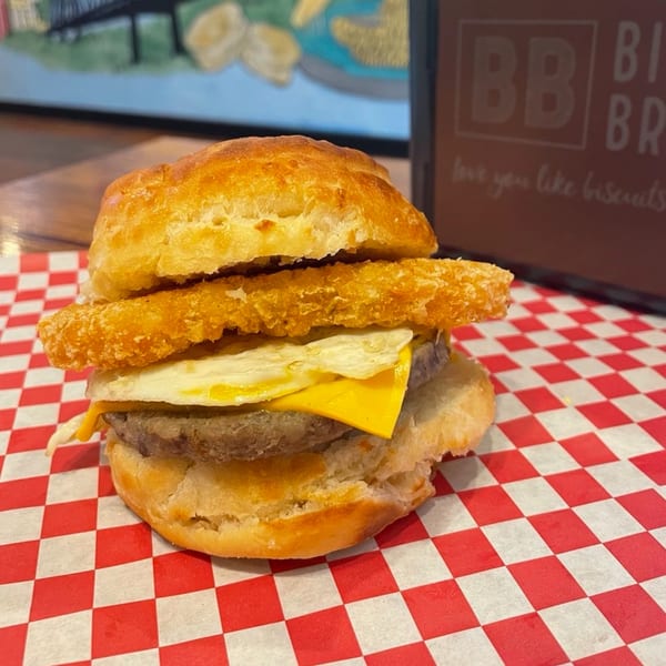A Biscuit Bros breakfast sandwich with sausage, egg, cheese, and a hash brown on a red checkered paper.