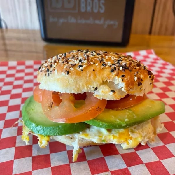 Everything bagel sandwich with egg, avocado, and tomato on red and white checkered paper.