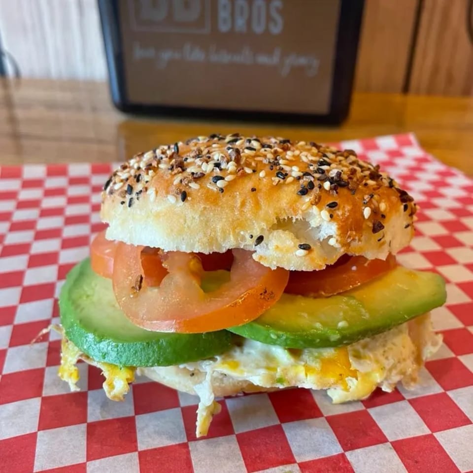 Everything bagel sandwich with egg, avocado, and tomato on red and white checkered paper.